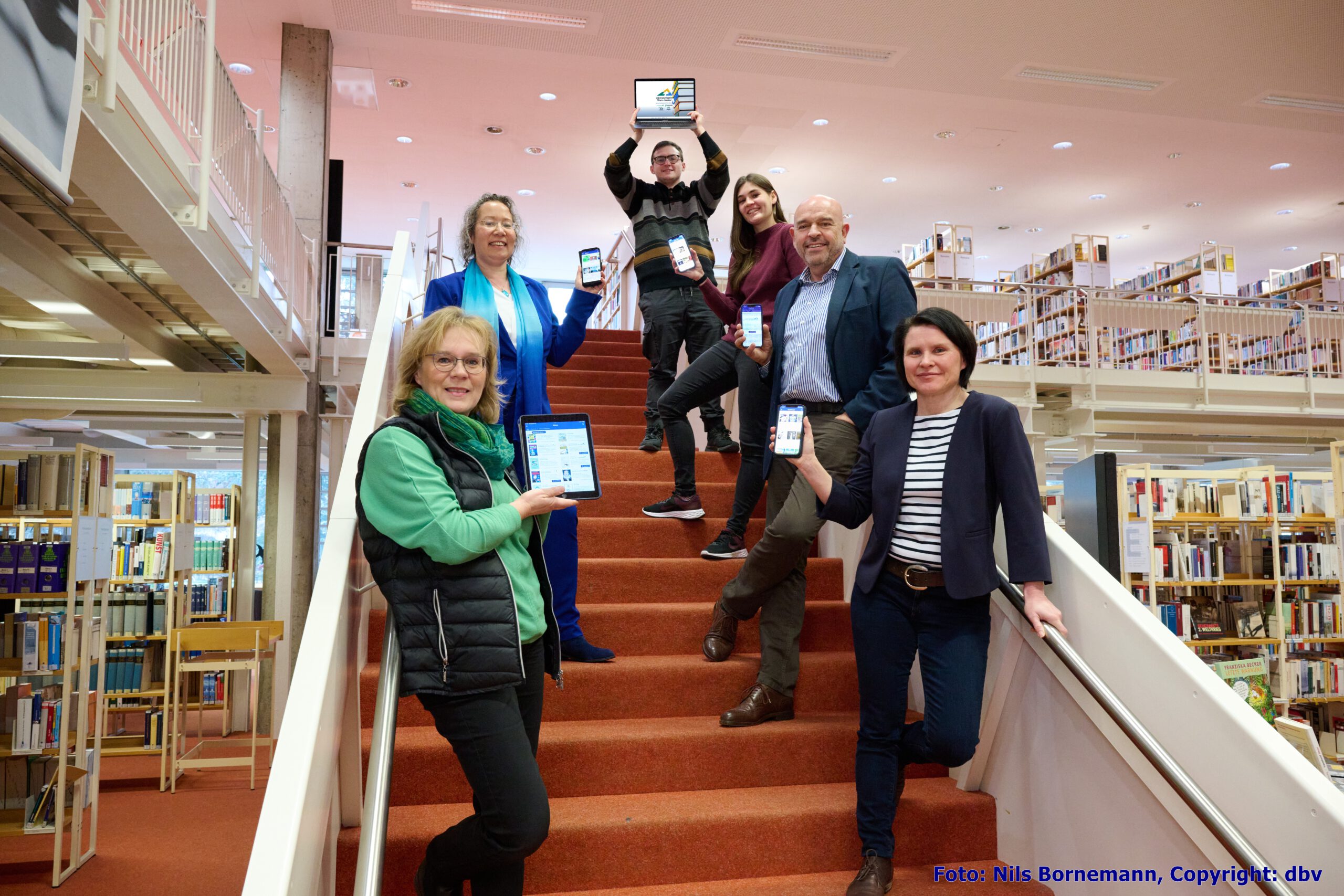 Gruppe auf Treppe in der Bücherei alle halten Smartphone oder eReader oder Notebook mit metropolbib in der Hand